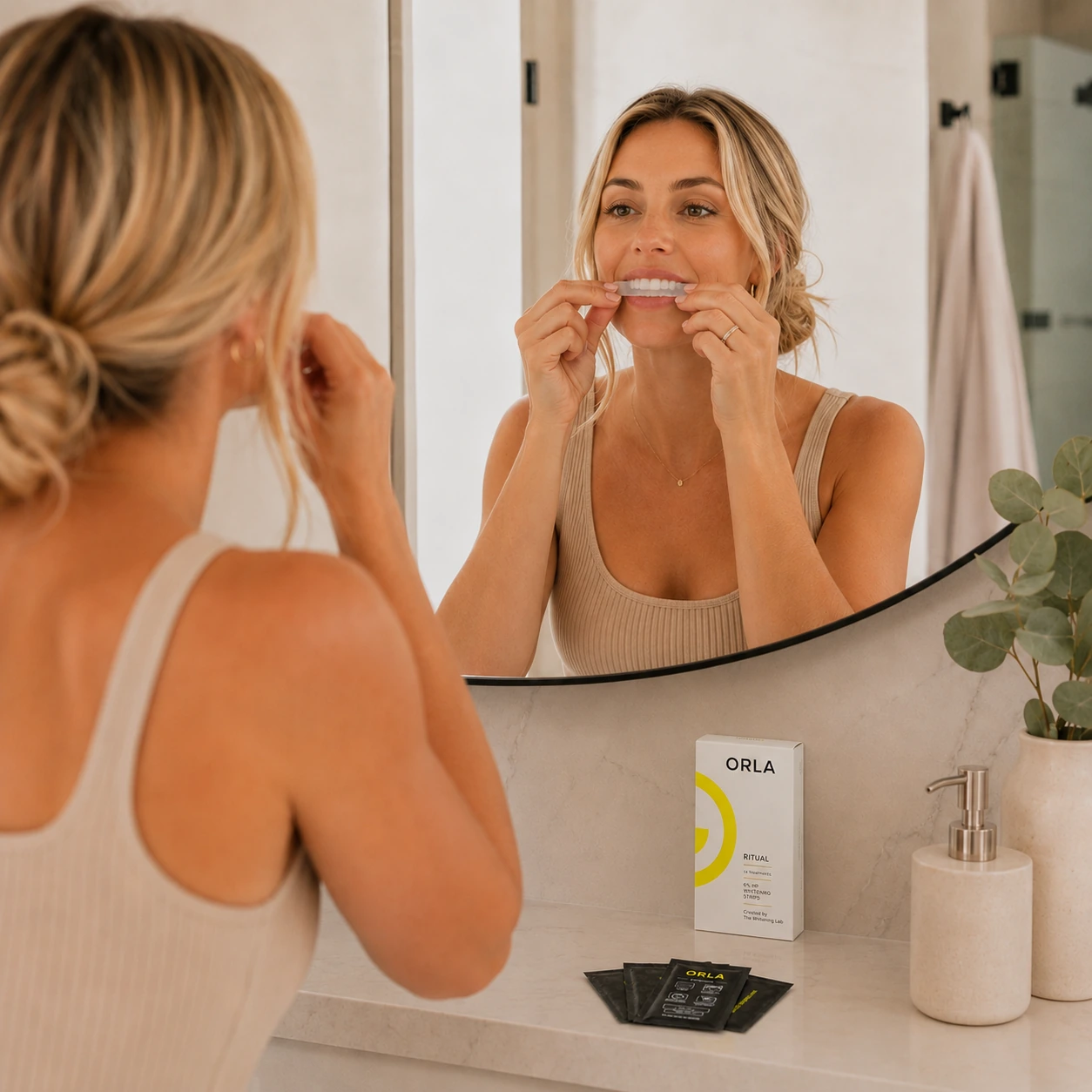 Woman applying ORLA™ whitening strips in front of a mirror with the ORLA™ Ritual box on the counter